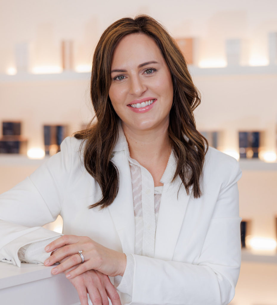 Kate Shneyder, Clinical Operations & Practice Development Consultant at glo MD Aesthetics and Wellness stands in front of product wall.